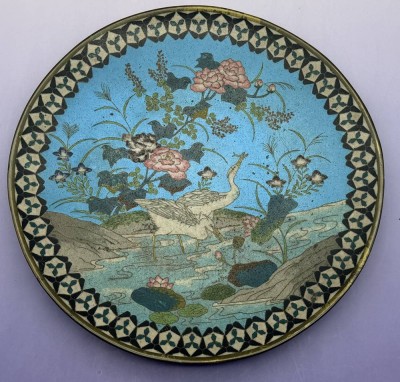Auktion 373<br>Cloisonne Teller der Meiji-Ära (1868-1912) mit Cloisonné-Emaillierung auf Metallgrund (Kupfer oder Bronze).Natur- und Uferszene mit Reihern/Wasservögeln,   auf hellblauem Grund. Ornamentaler geometrischer Bordürenrand in Schwarz und Weiß mit Metalldrahteinlagen. D-30,5 cm [1]